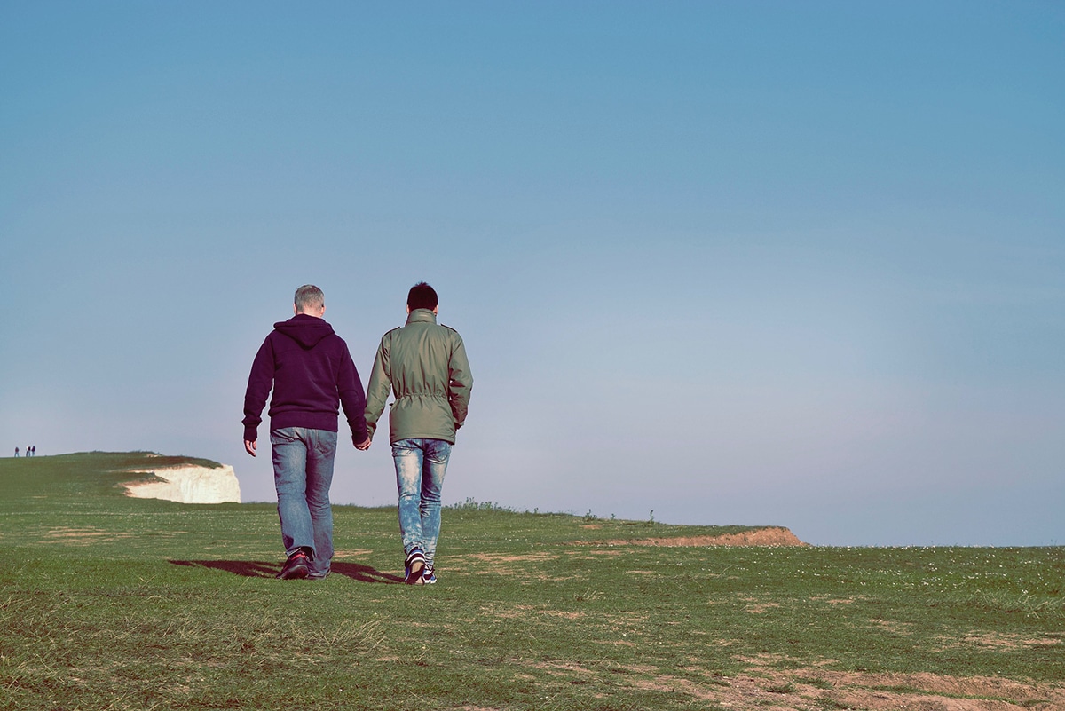 Two people holding hands while walking on a grassy field by a cliff