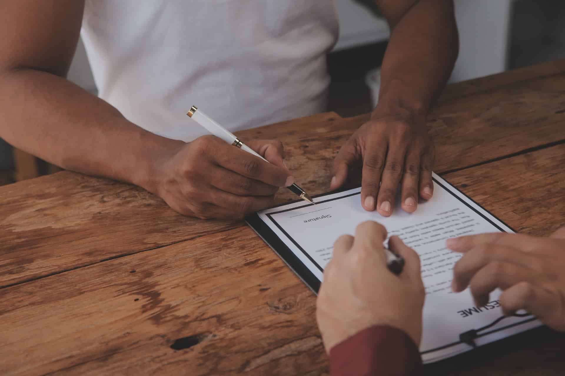 Two individuals signing a document on a clipboard at a rustic wooden table