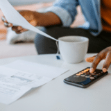 young man sitting at a table with a white coffee mug using his phone as a calculator to work on paperwork