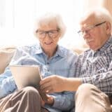 elderly couple sitting on a couch, looking at a tablet together with natural light shining through the window